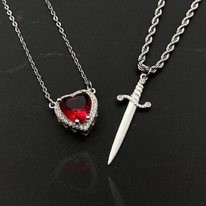 Op de afbeelding: Twee zilveren kettingen. Eén heeft een hartvormige rode edelsteen hanger met een halo van kleine heldere stenen. De andere heeft een zwaard hanger. Beide kettingen liggen op een zwart oppervlak.