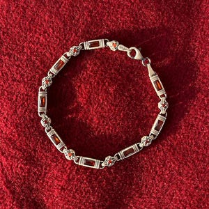 Peut inclure: Un bracelet en argent composé d'une alternance de pierres précieuses rectangulaires rouges et de maillons en argent ornés. Le bracelet est doté d'un fermoir mousqueton et présente une série de petites pierres rouge foncé serties.