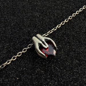 Könnte beinhalten: Halskette mit silberfarbenem Anhänger, der Hände zeigt, die einen herzförmigen, tiefroten Edelstein halten. Die Kette ist filigran, der Anhänger detailliert. Der Hintergrund ist eine dunkle, strukturierte Oberfläche.