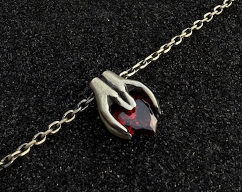 May include: A silver-toned necklace featuring a pendant of two hands cradling a heart-shaped, deep red gemstone. The chain is delicate, and the pendant has intricate details. The background is a dark, textured surface.
