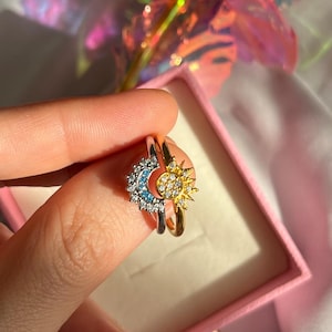 Peut inclure: Deux bagues superposées, une argentée et une dorée. La bague argentée présente un croissant de lune orné de pierres bleues et blanches. La bague dorée a un motif de soleil avec des pierres jaunes. Les bagues sont dans une boîte rose.