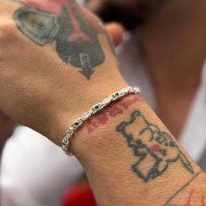 May include: A silver chain bracelet featuring a series of small, round, dark green gemstones set at regular intervals within the metallic links. The bracelet has a polished finish and a flexible, articulated design that wraps around the wrist.