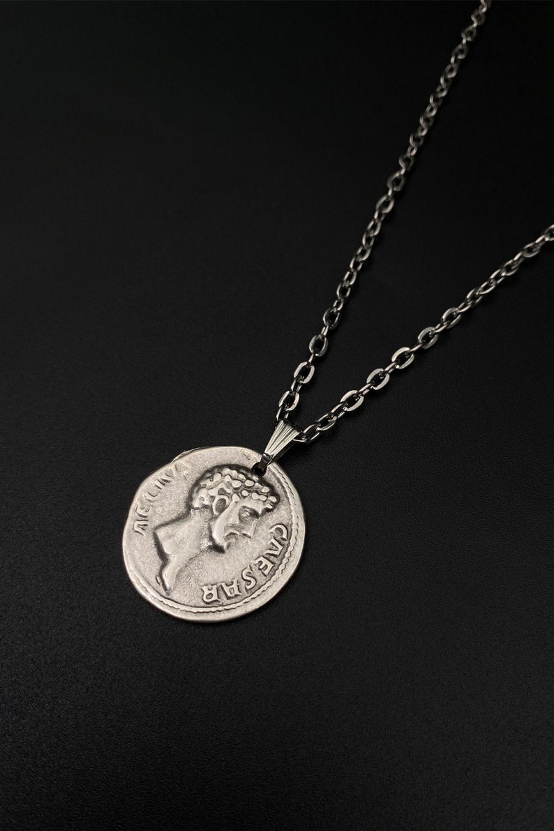 May include: A silver chain necklace with a silver coin pendant featuring a profile of Julius Caesar with the inscription "CAESAR" and "AET.CN.V" on the coin.