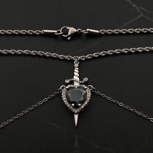 Op de afbeelding: Een zilveren ketting met een zwarte hart hanger met een zilveren dolk en heldere kristallen accenten.