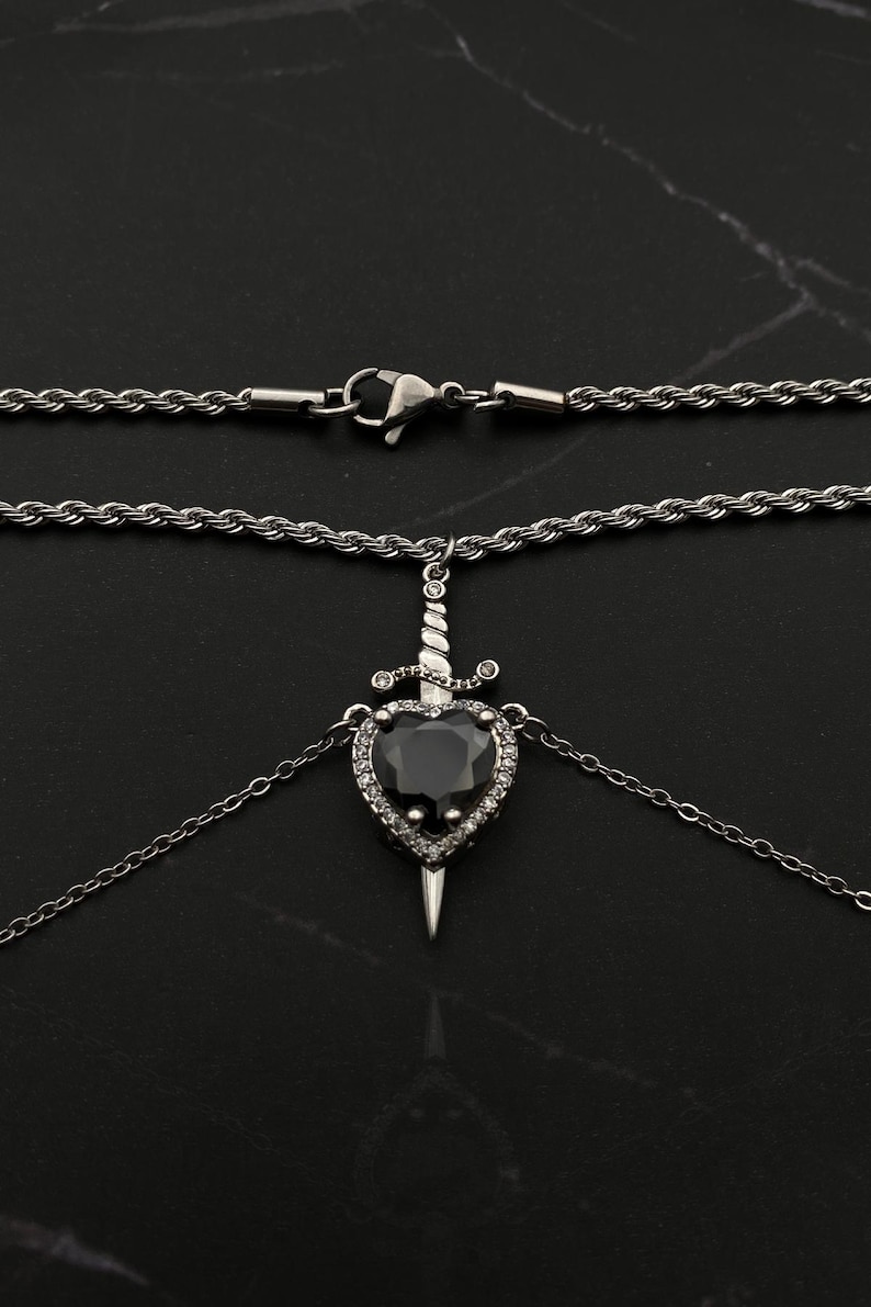Op de afbeelding: Een zilveren ketting met een zwarte hart hanger met een zilveren dolk en heldere kristallen accenten.