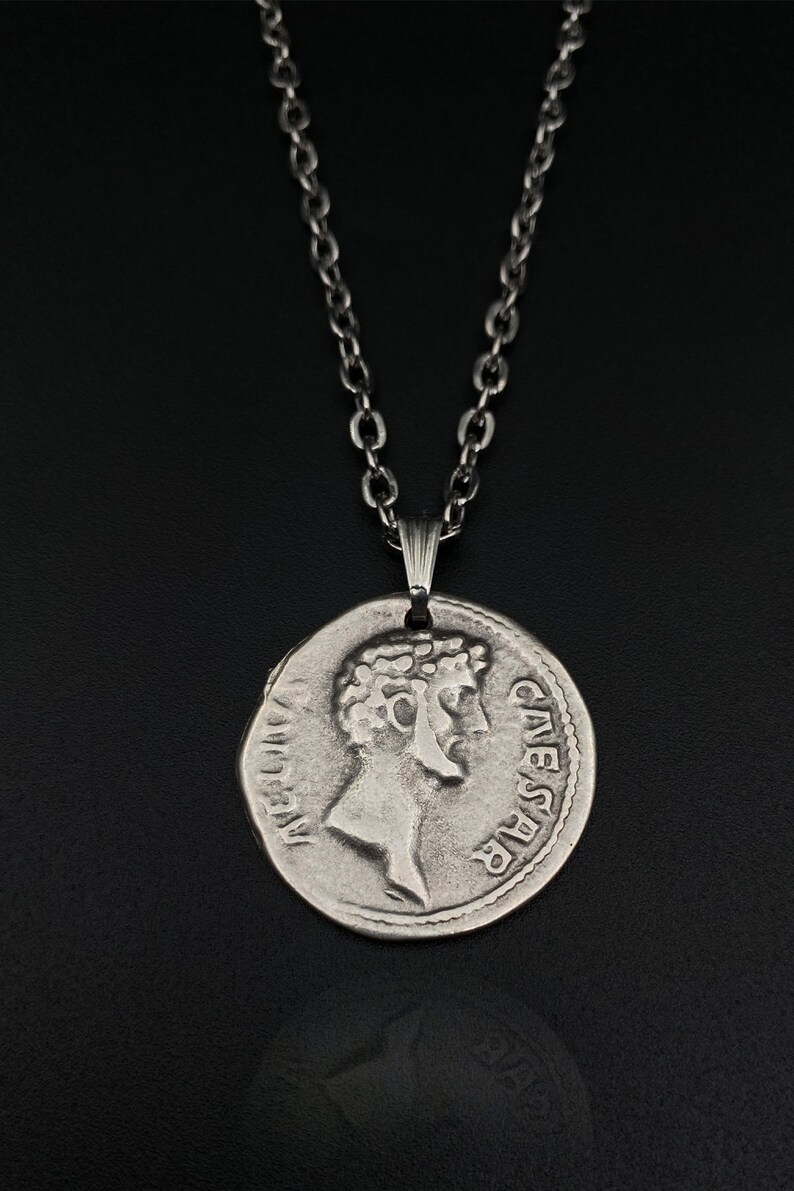 May include: A silver chain necklace with a silver coin pendant featuring a profile of Julius Caesar with the inscription "CAESAR" on the coin.
