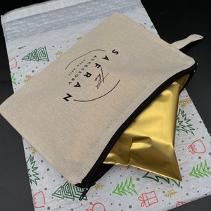 K&ouml;nnte beinhalten: Eine wei&szlig;e Canvas-Rei&szlig;verschlusstasche mit einem schwarzen Rei&szlig;verschluss und einem schwarzen Kreislogo, auf dem "The Safran Accessory" in wei&szlig;er Schrift steht. Die Tasche ist teilweise ge&ouml;ffnet und eine goldene Folient&uuml;te befindet sich im Inneren. Die Tasche befindet sich auf einem wei&szlig;en Hintergrund mit einem sich wiederholenden Muster aus gr&uuml;nen Weihnachtsb&auml;umen und roten Geschenken.