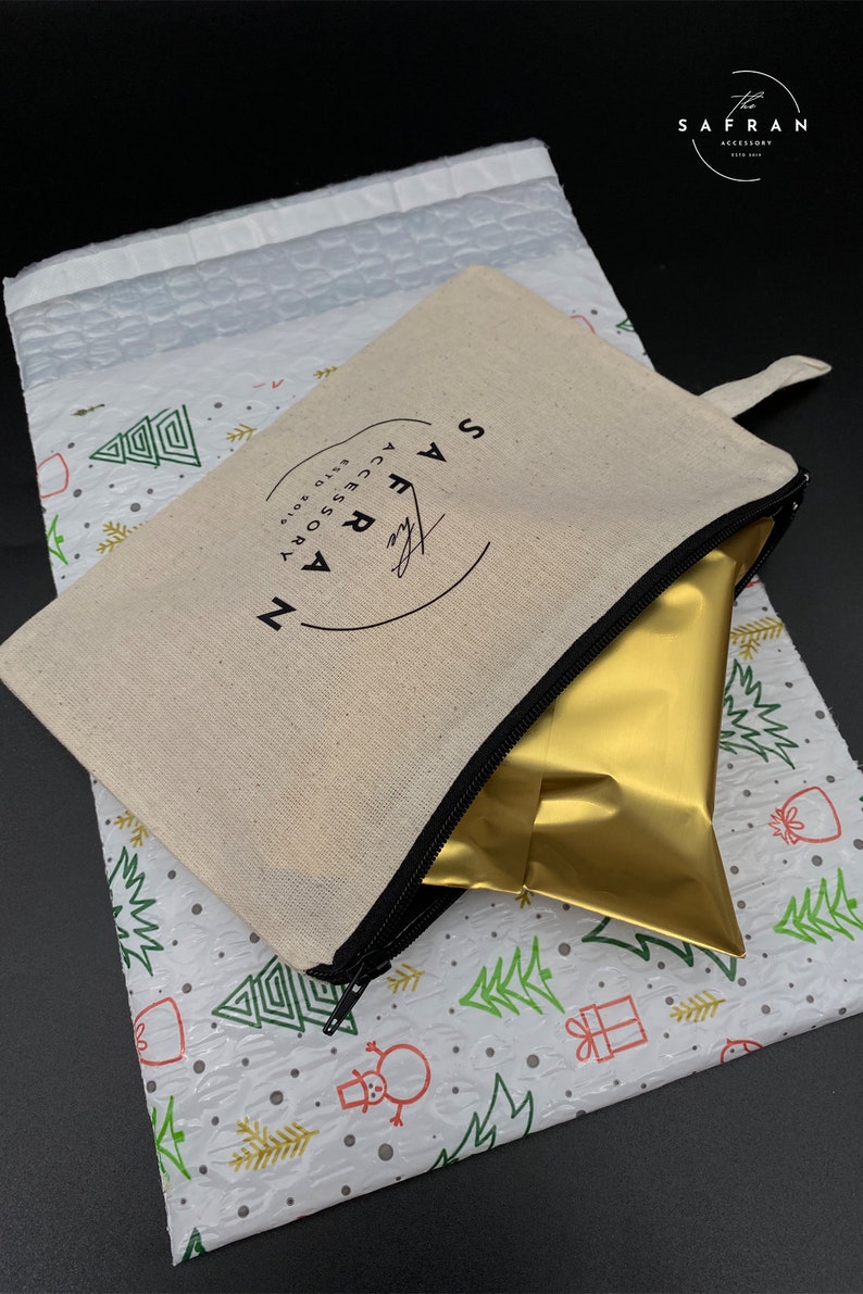 K&ouml;nnte beinhalten: Eine wei&szlig;e Canvas-Rei&szlig;verschlusstasche mit einem schwarzen Rei&szlig;verschluss und einem schwarzen Kreislogo, auf dem "The Safran Accessory" in wei&szlig;er Schrift steht. Die Tasche ist teilweise ge&ouml;ffnet und eine goldene Folient&uuml;te befindet sich im Inneren. Die Tasche befindet sich auf einem wei&szlig;en Hintergrund mit einem sich wiederholenden Muster aus gr&uuml;nen Weihnachtsb&auml;umen und roten Geschenken.