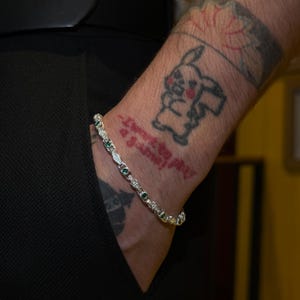 May include: Silver bracelet with emerald and clear stone accents. The bracelet is worn on a wrist with tattoos, including a Pikachu design. The person is wearing a black outfit.