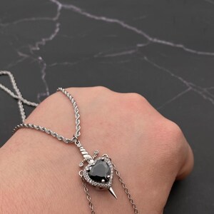Op de afbeelding: Een zilveren ketting met een zwarte hartvormige hanger met een zilveren dolk. Het hart is omgeven door kleine, heldere stenen.