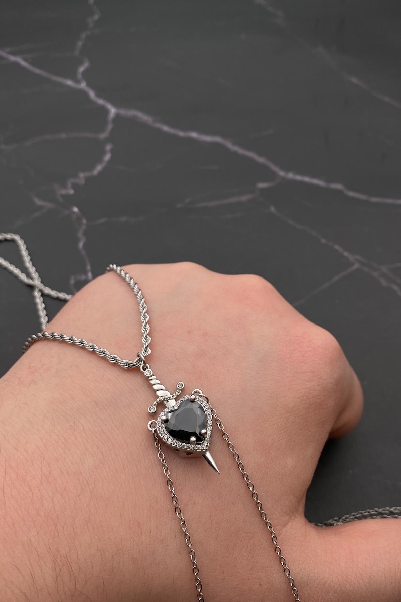 Op de afbeelding: Een zilveren ketting met een zwarte hartvormige hanger met een zilveren dolk. Het hart is omgeven door kleine, heldere stenen.