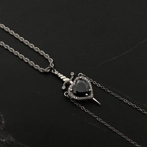 Op de afbeelding: Een zilveren ketting met een zwarte hart hanger met een zilveren dolk die door het midden gaat. Het hart is omgeven door kleine heldere stenen.
