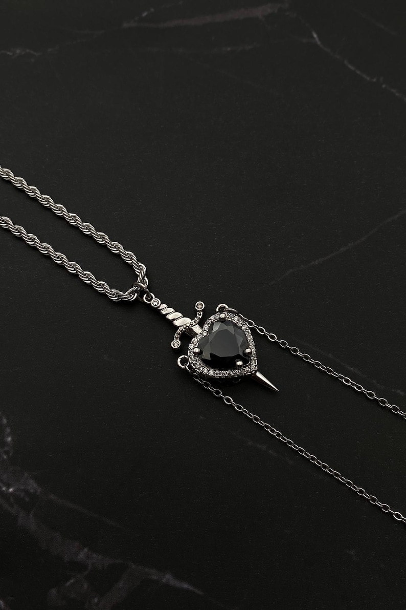 Op de afbeelding: Een zilveren ketting met een zwarte hart hanger met een zilveren dolk die door het midden gaat. Het hart is omgeven door kleine heldere stenen.