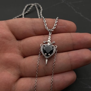 Op de afbeelding: Een zilveren ketting met een zwarte hart hanger met een zilveren dolk die door het midden steekt. Het hart is omgeven door kleine heldere stenen.