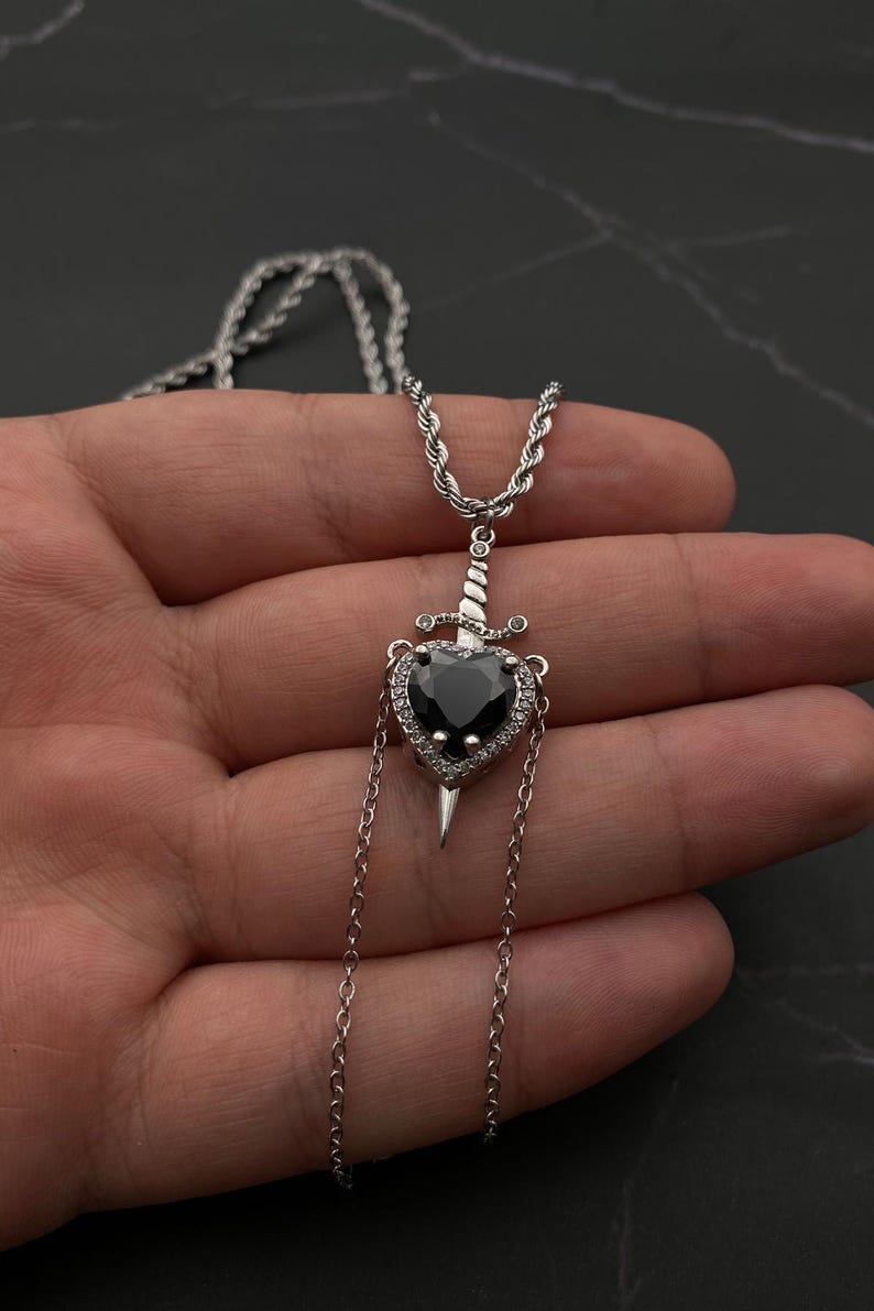 Op de afbeelding: Een zilveren ketting met een zwarte hart hanger met een zilveren dolk die door het midden steekt. Het hart is omgeven door kleine heldere stenen.