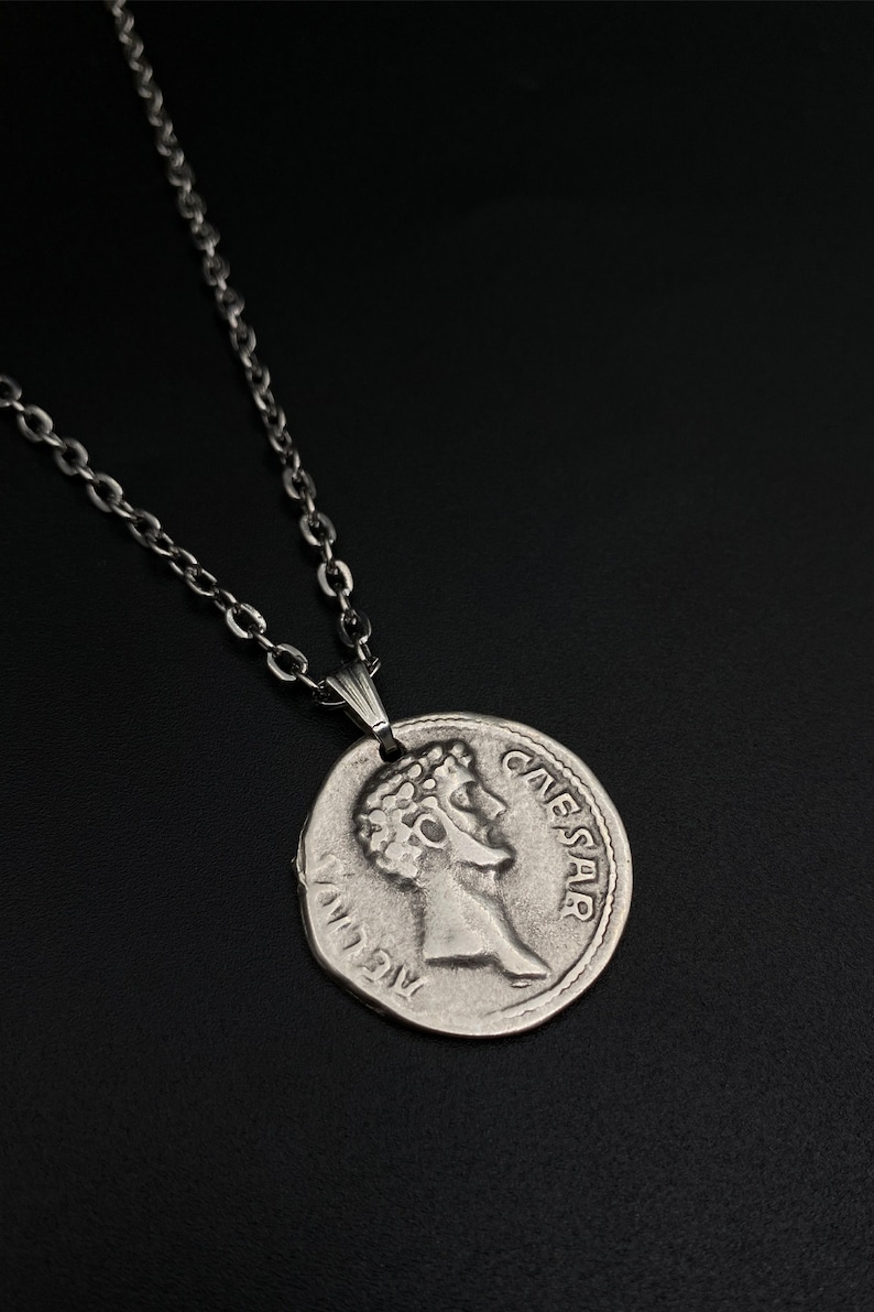 May include: Silver chain necklace with a silver coin pendant featuring a profile of Julius Caesar and the inscription "CAESAR".