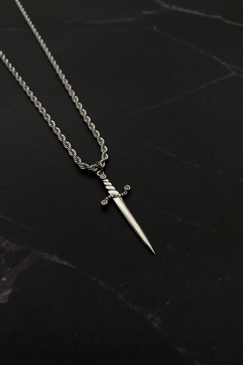 Op de afbeelding: Een zilveren touwketting met een zilveren zwaardhanger. Het zwaard heeft een gedraaid ontwerp en kleine heldere stenen op het handvat.