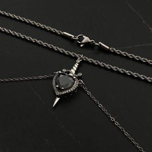 Op de afbeelding: Een zilveren ketting met een zwarte hartvormige hanger met een zilveren dolk. De hanger is versierd met kleine heldere stenen. De ketting heeft een touwketting en een karabijnhaak.