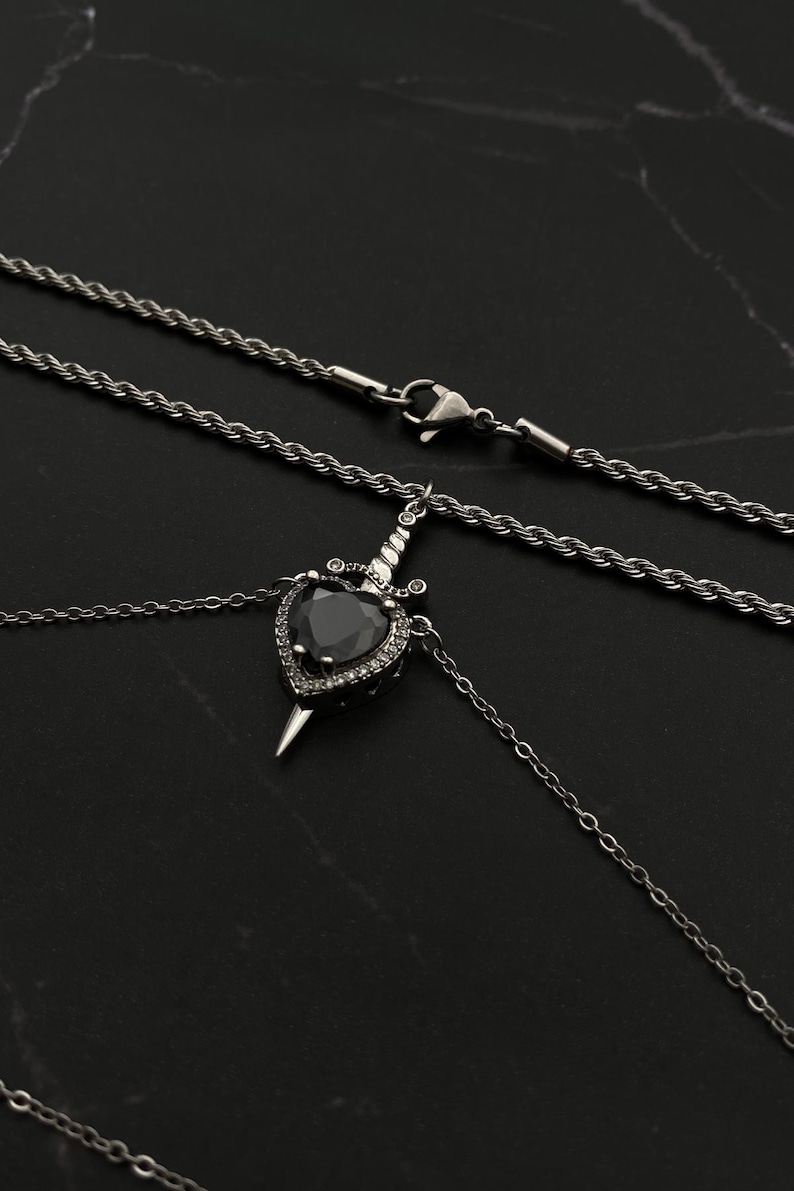 Op de afbeelding: Een zilveren ketting met een zwarte hartvormige hanger met een zilveren dolk. De hanger is versierd met kleine heldere stenen. De ketting heeft een touwketting en een karabijnhaak.