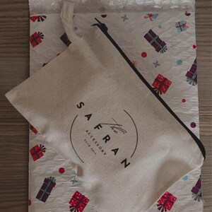 Op de afbeelding: Een witte canvas tas met een zwarte rits en een zwart logo met de tekst "The Safran Accessory Est. 2017" op een witte achtergrond met een patroon van rode en paarse cadeaudozen.