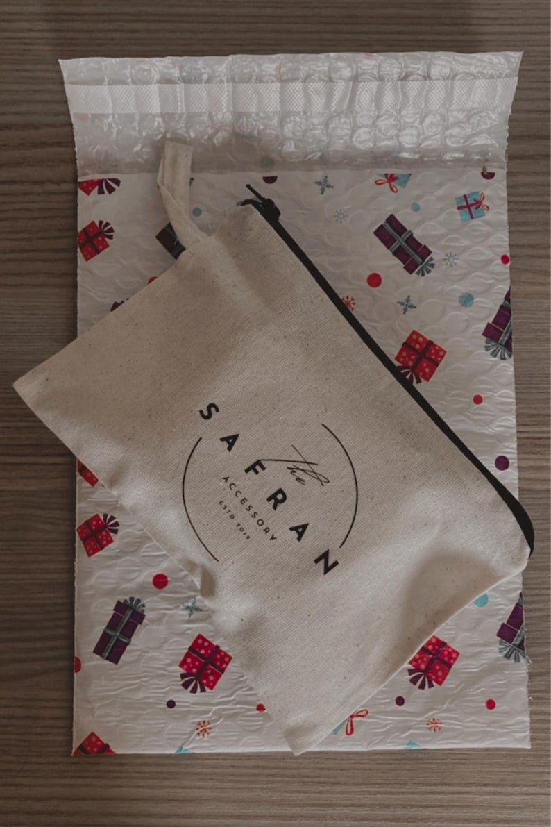 Op de afbeelding: Een witte canvas tas met een zwarte rits en een zwart logo met de tekst "The Safran Accessory Est. 2017" op een witte achtergrond met een patroon van rode en paarse cadeaudozen.