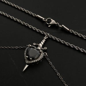 Op de afbeelding: Een zilveren ketting met een zwarte hartvormige hanger met een zilveren dolk. Het hart is versierd met kleinere, heldere stenen.