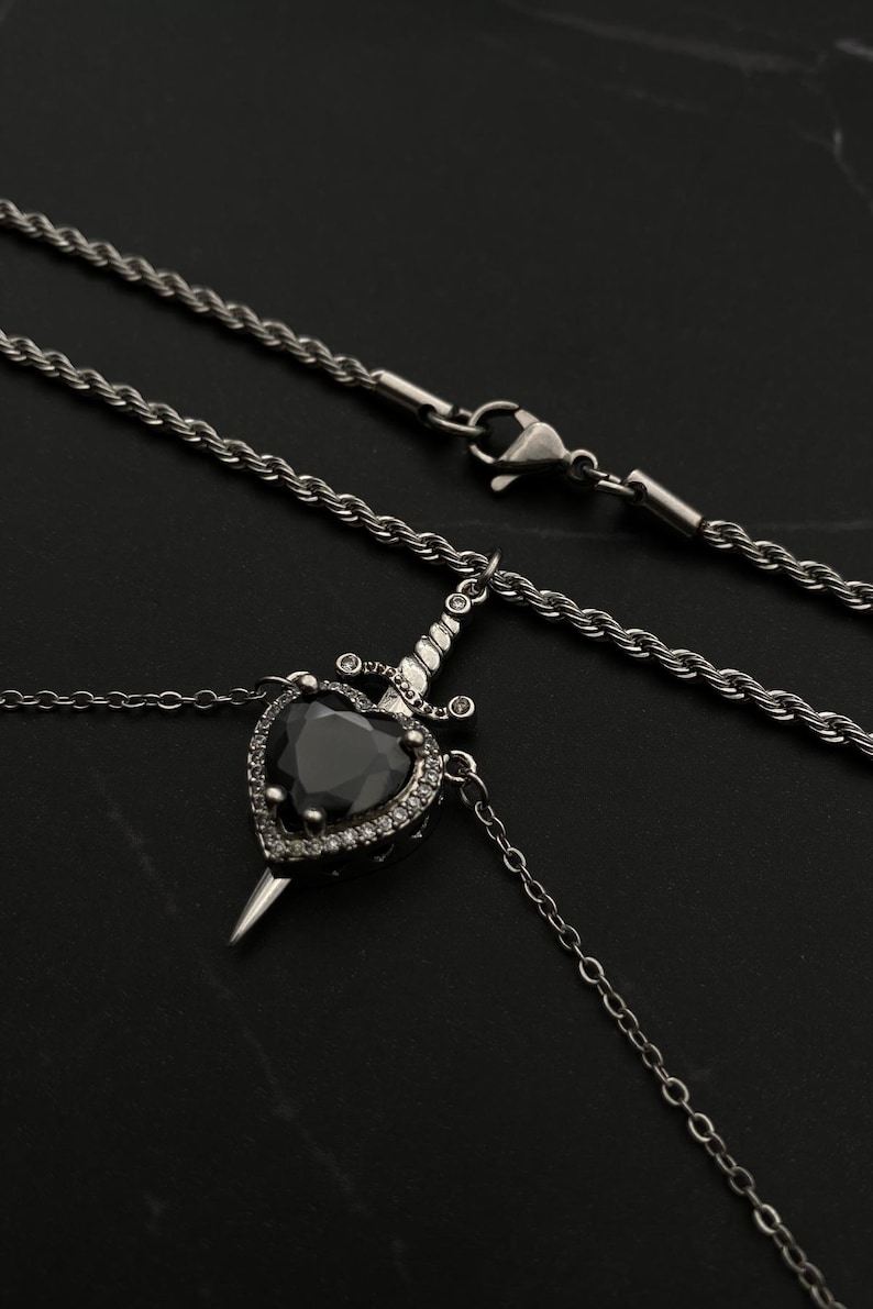 Op de afbeelding: Een zilveren ketting met een zwarte hartvormige hanger met een zilveren dolk. Het hart is versierd met kleinere, heldere stenen.