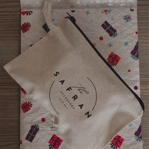 K&ouml;nnte beinhalten: Eine wei&szlig;e Stofftasche mit einem schwarzen Rei&szlig;verschluss und dem aufgedruckten Text "The Saffron Accessory Est'd 2015" in Schwarz.