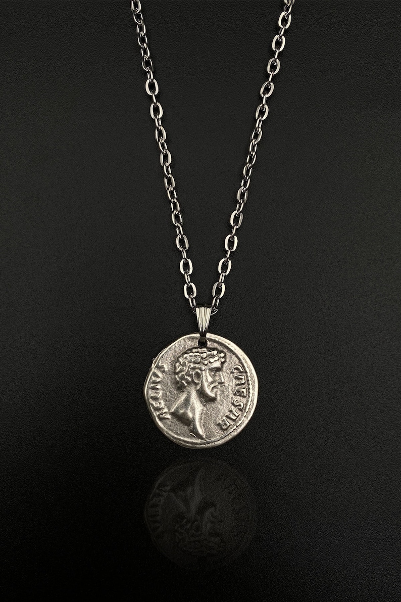 May include: A silver chain necklace with a silver coin pendant featuring a profile of a man's face. The coin has the words "IMP CAESAR" and "CIVES SPQR" inscribed on it.
