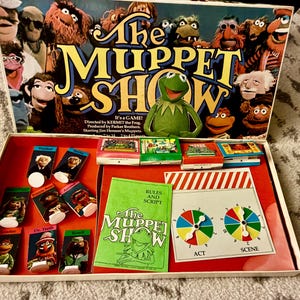 May include: The Muppet Show board game featuring a box lid with various Muppet characters, a green Rules and Script booklet, eight character standees, four small game boxes, and a game board with two colorful numbered spinners.