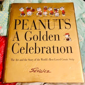 May include: A gold-colored book titled "PEANUTS A Golden Celebration." The cover features illustrations of Peanuts characters and the text "The Art and the Story of the World's Best-Loved Comic Strip." The author's signature, SCHULZ, is at the bottom.