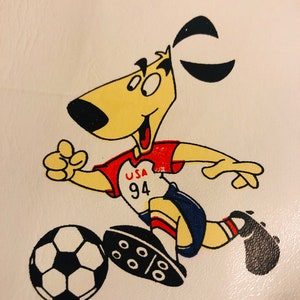 Puede incluir: Un perro de dibujos animados con uniforme de f&uacute;tbol, pateando un bal&oacute;n de f&uacute;tbol. El perro es amarillo con orejas negras y una nariz negra. El uniforme es rojo y blanco con "USA 94" en el pecho. El bal&oacute;n de f&uacute;tbol es blanco y negro.