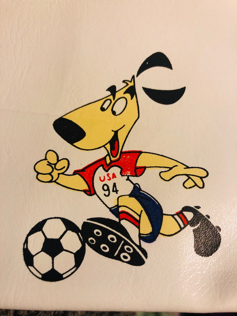 Puede incluir: Un perro de dibujos animados con uniforme de f&uacute;tbol, pateando un bal&oacute;n de f&uacute;tbol. El perro es amarillo con orejas negras y una nariz negra. El uniforme es rojo y blanco con "USA 94" en el pecho. El bal&oacute;n de f&uacute;tbol es blanco y negro.