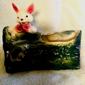 May include: A ceramic planter shaped like a log with a small rabbit figurine sitting on top. The rabbit is white with pink ears and a red bow. The planter is green and brown with a textured surface.