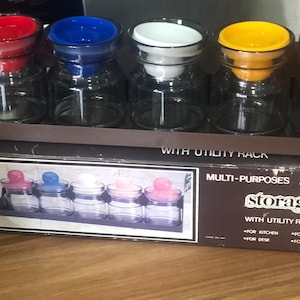 May include: A set of five clear glass storage jars with colorful lids in red, blue, white, yellow, and orange. The jars are displayed in a brown cardboard box with the text "storage jar".