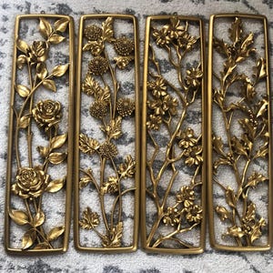 May include: Four rectangular gold-colored wall panels with floral designs. Each panel features a different arrangement of flowers and foliage, including roses, chrysanthemums, and other blossoms. The panels are framed and appear to be made of metal.
