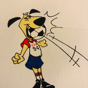 Puede incluir: Un perro de dibujos animados con un uniforme de f&uacute;tbol rojo, blanco y azul, con el texto "USA 94" en la camiseta. El perro sonr&iacute;e y parece estar pateando un bal&oacute;n de f&uacute;tbol. L&iacute;neas negras indican la trayectoria del bal&oacute;n.