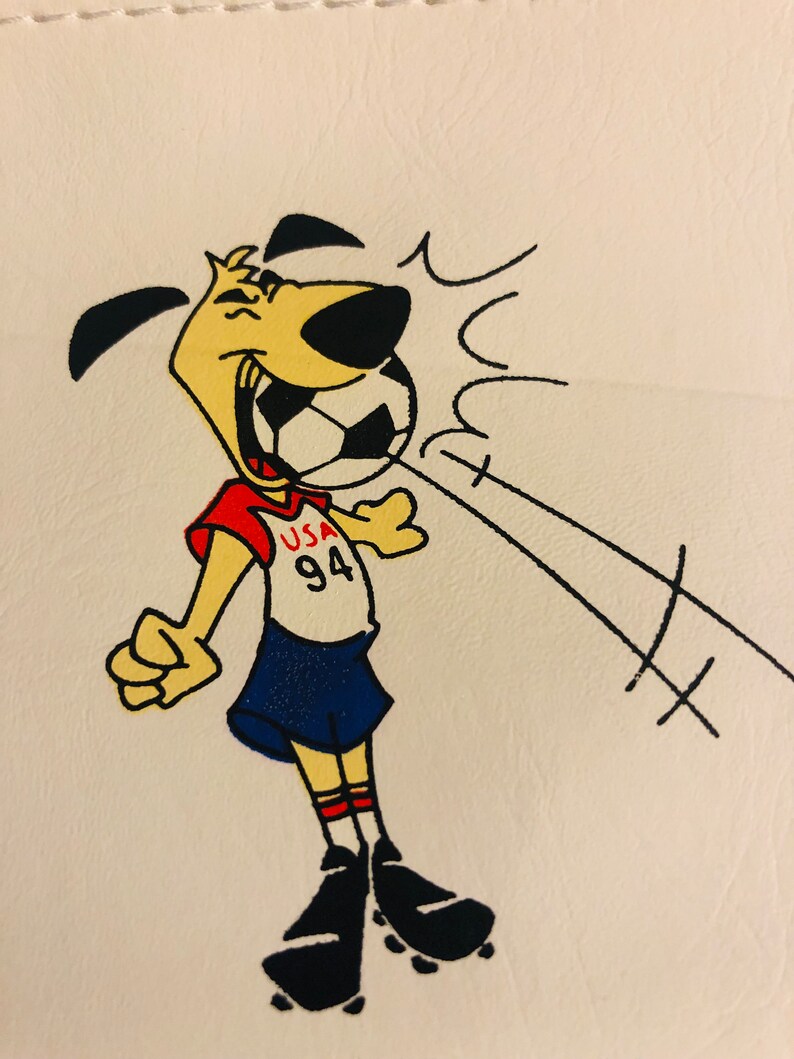 Puede incluir: Un perro de dibujos animados con un uniforme de f&uacute;tbol rojo, blanco y azul, con el texto "USA 94" en la camiseta. El perro sonr&iacute;e y parece estar pateando un bal&oacute;n de f&uacute;tbol. L&iacute;neas negras indican la trayectoria del bal&oacute;n.