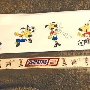 Puede incluir: Estuche blanco con perros de dibujos animados jugando al f&uacute;tbol. Los perros visten uniformes rojos y azules. Debajo del estuche hay una regla con el logo de Snickers y m&aacute;s ilustraciones de perros futbolistas.