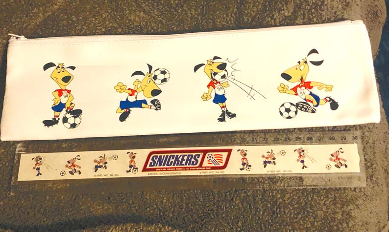 Puede incluir: Estuche blanco con perros de dibujos animados jugando al f&uacute;tbol. Los perros visten uniformes rojos y azules. Debajo del estuche hay una regla con el logo de Snickers y m&aacute;s ilustraciones de perros futbolistas.