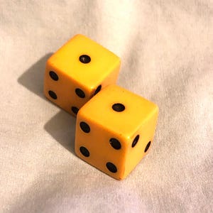 May include: Two yellow dice with black dots, resting on a white surface. The dice are cube-shaped, with the dots arranged in the standard patterns for dice. The dice are a warm yellow color, and the dots are black.