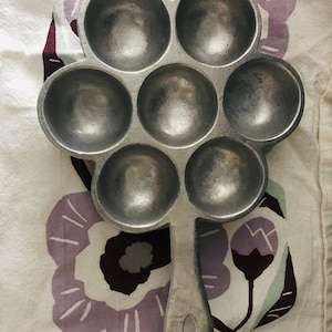 May include: A silver-colored cast iron pan with seven round indentations for baking. The pan has a long handle and is shaped like a flower.