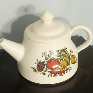 May include: A white ceramic teapot with a vegetable design. The teapot has a lid and a handle. The vegetables are painted in shades of green, red, yellow, and brown.