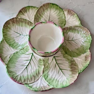 May include: A ceramic serving platter shaped like a large cabbage leaf with six distinct sections surrounding a central bowl. The dish features a cream-colored base with hand-painted green and pink edges, detailed with embossed leaf veins throughout.