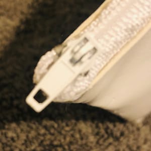 Puede incluir: Primer plano de un tirador de cremallera y cinta de cremallera blancos. El tirador de la cremallera es rectangular con un agujero cuadrado. La cinta de la cremallera es blanca y parece estar hecha de un material tejido. El fondo es borroso y texturizado, de color gris.