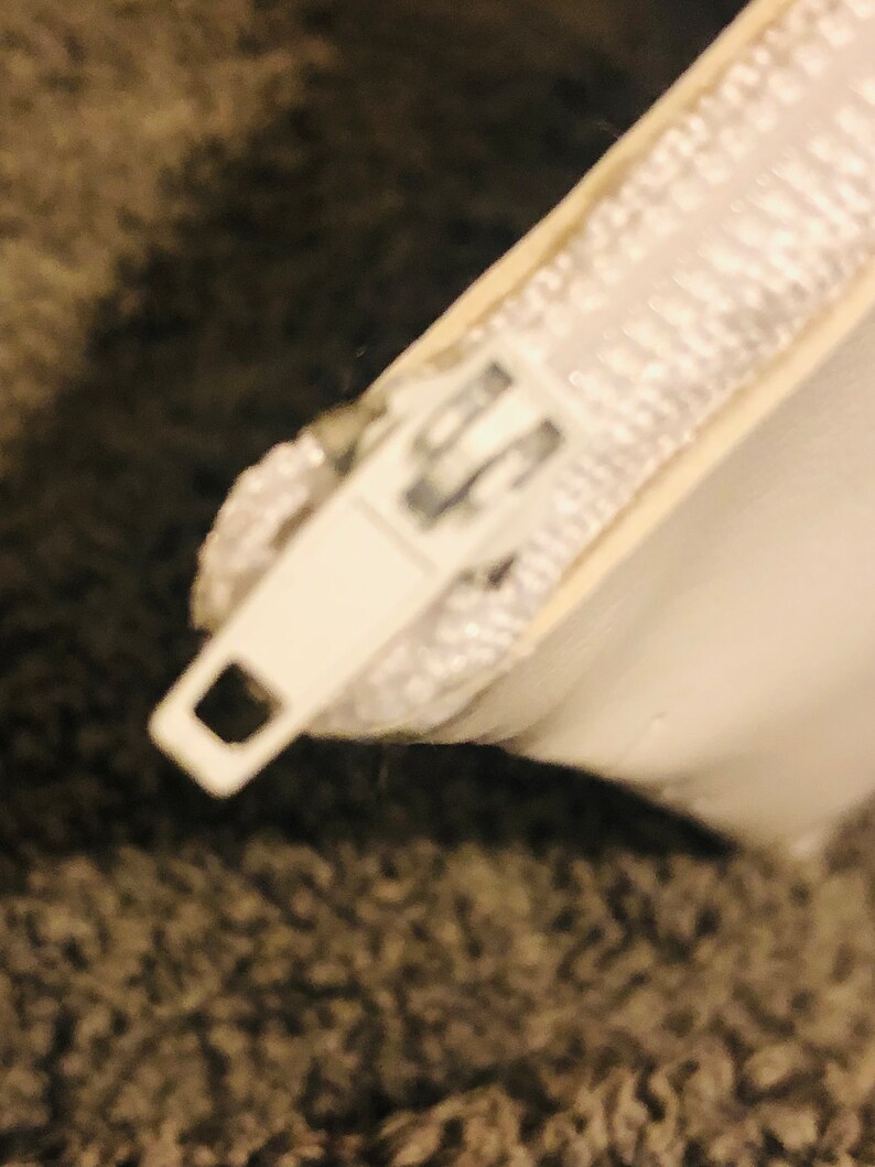 Puede incluir: Primer plano de un tirador de cremallera y cinta de cremallera blancos. El tirador de la cremallera es rectangular con un agujero cuadrado. La cinta de la cremallera es blanca y parece estar hecha de un material tejido. El fondo es borroso y texturizado, de color gris.