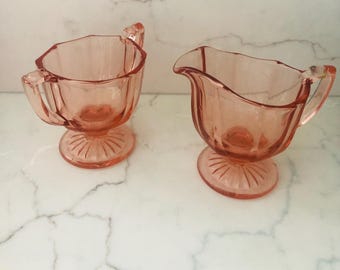May include: A vintage pink glass sugar bowl and creamer set with a fluted design. The set is on a white marble surface.
