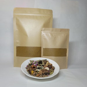 May include: Two brown paper bags with a zipper closure, one larger than the other, both filled with brown powder. A white bowl with a variety of spices and herbs, including star anise, cloves, and bay leaves.