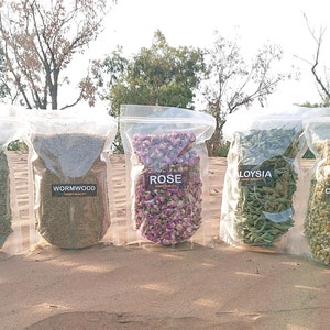 Op de afbeelding: Zes doorzichtige plastic zakken gevuld met gedroogde kruiden en bloemen. De zakken zijn gelabeld met de namen van de kruiden en bloemen: Tijm, Rozemarijn, Absint, Roos,  Alloysa, Kamille en Lavendel.