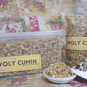 Op de afbeelding: Twee containers met gedroogde, lichtbruine, hele komijnzaadjes. De containers zijn gelabeld met "Woly Cumin Moroccan Product".